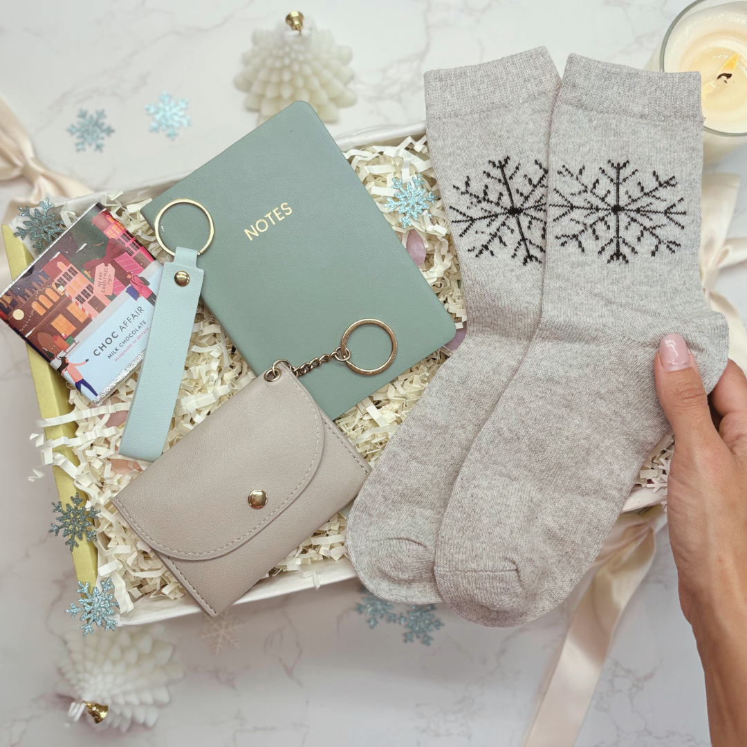 Gray socks with snowflake design held by a hand, next to a small beige purse and green notebook on a decorative surface.