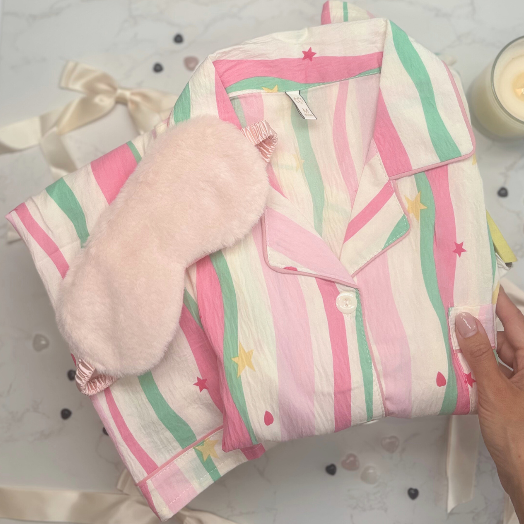 Folded pajama set with pink and green stripes, fluffy pink eye mask on a lemon yellow gift box. 