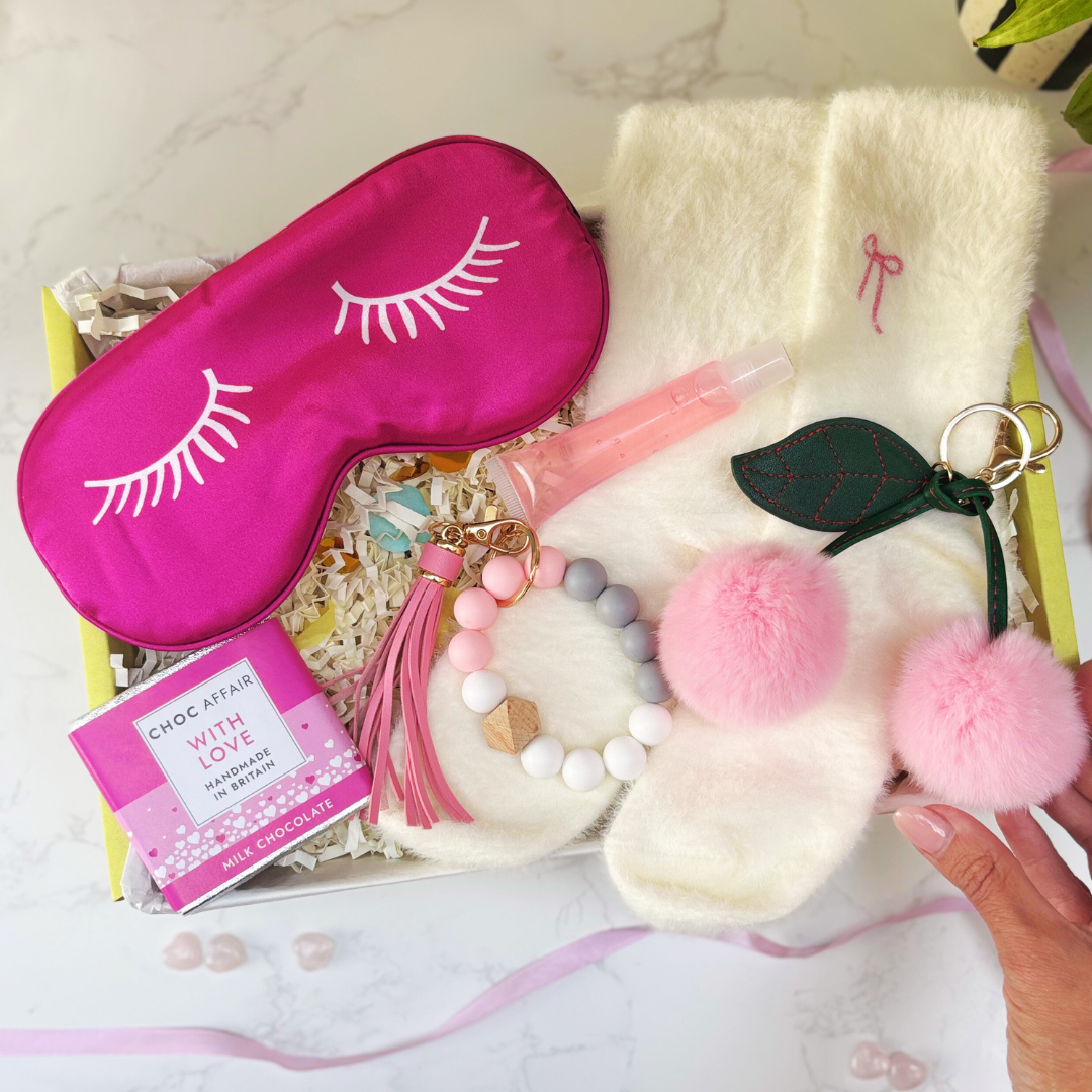 Pink sleep mask with eyelashes, fluffy white socks, lip balm and cherry keychain with pink pom-poms in a gift box.
