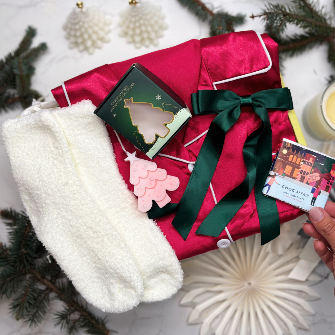 Satin feel festive deep pink pyjamas with white fluffy socks, christmas tree candle and satin green hair ribbon, held against a decorative giftbox.