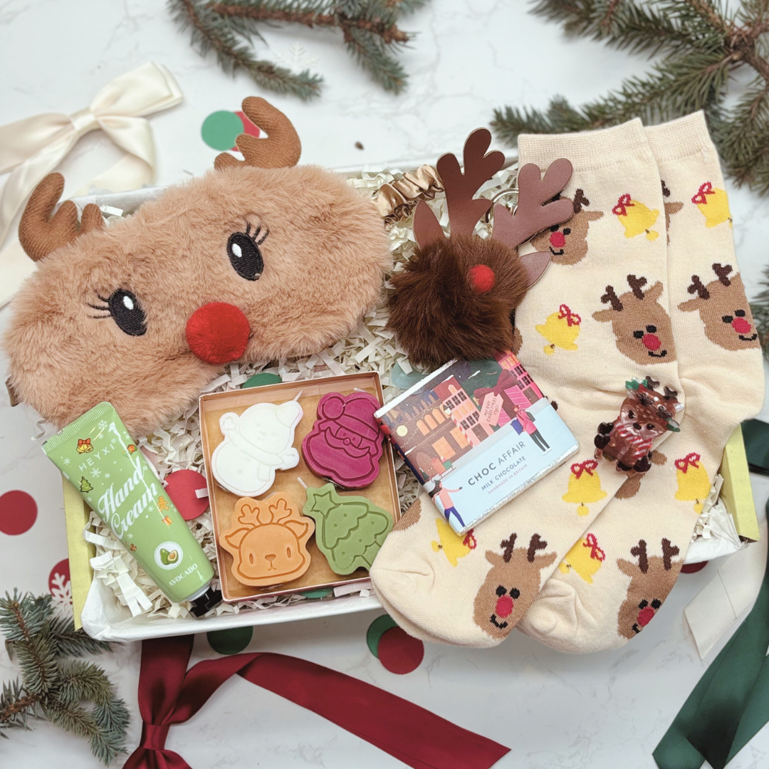 Christmas gift set with reindeer-themed items including a plush eye mask, candles, keyring, hairclip and socks on a yellow giftbox.