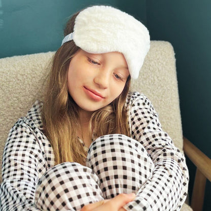 Person wearing gingham pjs and white fluffy eyemask sitting on a chair.