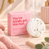 Pink shower steamer packaging box next to a round white shower steamer disc with visible green herb flecks, styled on a bathroom counter with a pink towel, gold faucet, and soft warm lighting.