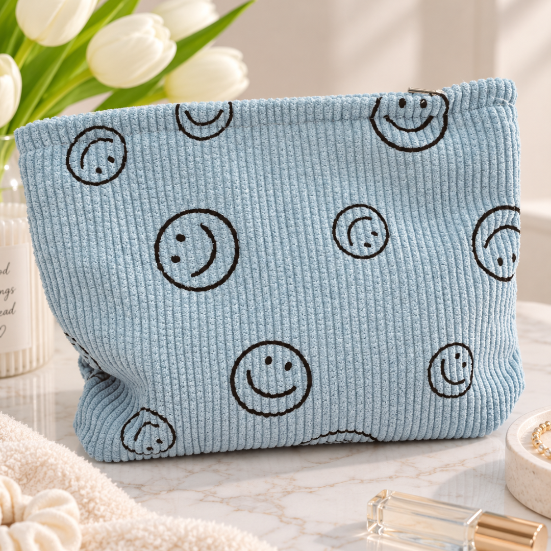 A light blue corduroy cosmetic pouch with a black smiley face pattern, styled on a marble surface with white tulips and skincare items in the background.