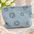 A light blue corduroy cosmetic pouch with a black smiley face pattern, styled on a marble surface with white tulips and skincare items in the background.