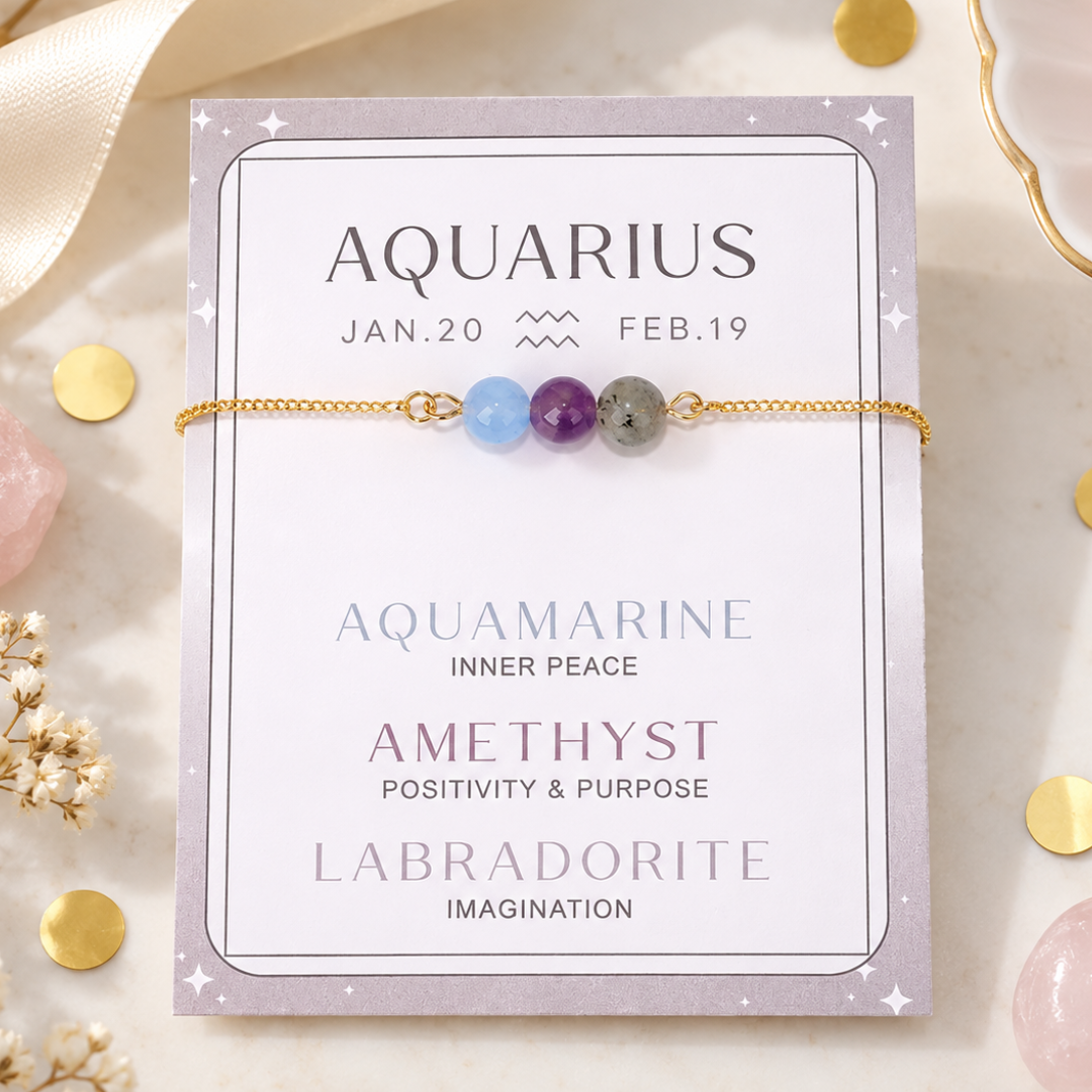A gold chain bracelet with three gemstone beads (aquamarine, amethyst, and labradorite) displayed on its lavender card packaging, styled on a marble surface with gold confetti and crystals.