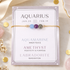 A gold chain bracelet with three gemstone beads (aquamarine, amethyst, and labradorite) displayed on its lavender card packaging, styled on a marble surface with gold confetti and crystals.
