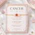 A gold beaded bracelet with pink, orange, and white rose quartz beads is displayed on its pink card packaging, which features astrological details for Cancer. The card is styled on a white marble surface with scattered rose quartz crystals and gold coins.