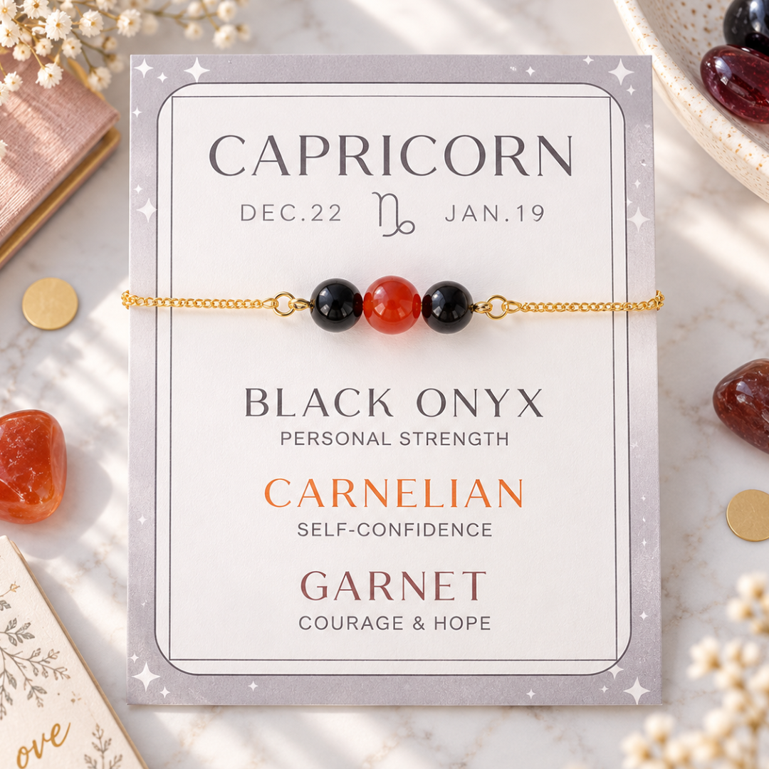 A gold chain bracelet with three beads (two black onyx, one red carnelian) displayed on a branded backing card that reads &