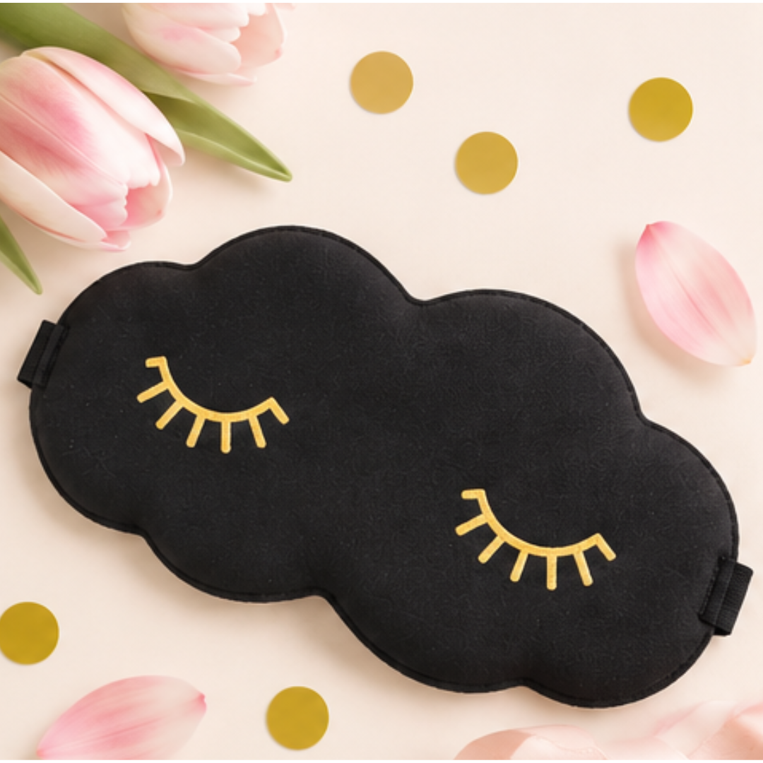 A black cloud-shaped sleep mask with gold embroidered closed-eye details, styled on a light beige background with pink tulips, petals, and gold confetti.