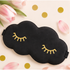 A black cloud-shaped sleep mask with gold embroidered closed-eye details, styled on a light beige background with pink tulips, petals, and gold confetti.