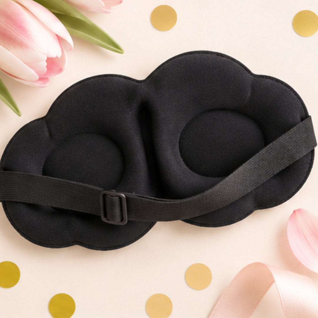 A black cloud-shaped eye mask with an adjustable strap and buckle, styled on a light beige background with pink tulips, gold confetti, and a pink ribbon.