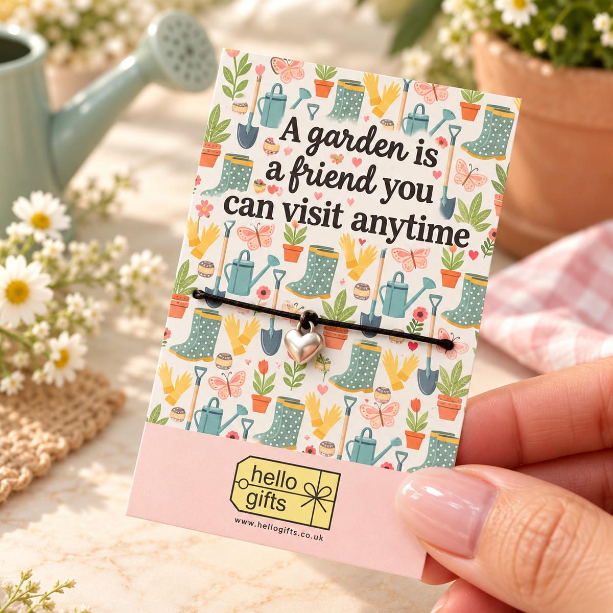 A silver heart charm on a black cord bracelet displayed on a decorative greeting card held by a hand. The card features gardening illustrations and the text &