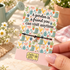 A silver heart charm on a black cord bracelet displayed on a decorative greeting card held by a hand. The card features gardening illustrations and the text &
