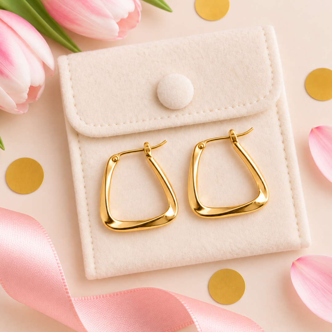 Gold hoop earrings with a modern wavy, open triangular design, displayed on a cream-colored velvet jewelry pouch. The pouch is styled on a light pink background with pink tulips, gold confetti, and a pink satin ribbon.