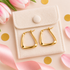 Gold hoop earrings with a modern wavy, open triangular design, displayed on a cream-colored velvet jewelry pouch. The pouch is styled on a light pink background with pink tulips, gold confetti, and a pink satin ribbon.