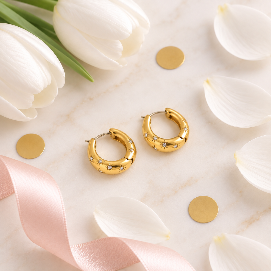 A pair of gold crescent-shaped hoop earrings with small star details, displayed on a light marble surface with white tulips, white petals, gold coins, and a pink ribbon.