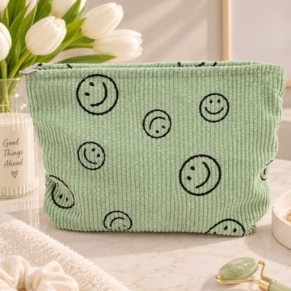 A mint green corduroy cosmetic pouch with black smiley face prints, styled on a marble surface with white tulips and skincare props in the background.