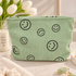 A mint green corduroy cosmetic pouch with black smiley face prints, styled on a marble surface with white tulips and skincare props in the background.