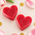 Two heart-shaped red soap bars arranged on a light pink background with decorative pink flower petals, gold confetti, and a green leaf.