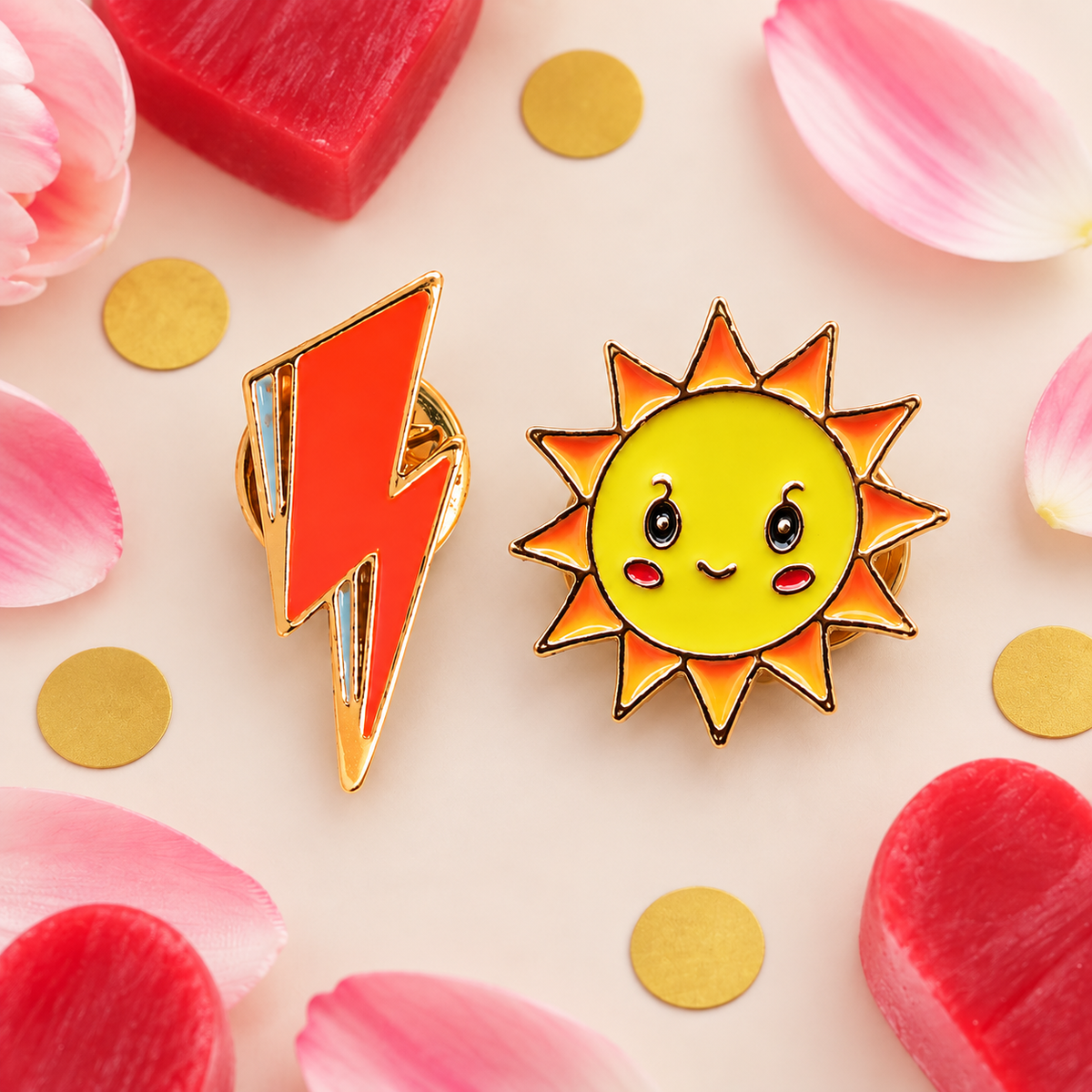 Two enamel pins, a red lightning bolt with gold trim and a smiling yellow sun with orange rays, displayed on a light pink background with pink flower petals and gold confetti.