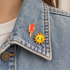 Two enamel pins, a red lightning bolt and a yellow smiling sun, pinned to the collar of a light blue denim jacket worn by a person.