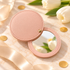 A round compact-style folding mirror with a light pink faux-leather cover, shown open on a beige marble-like surface with tulips, ribbon, and gold confetti props.