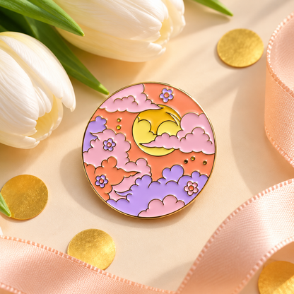 A round enamel pin featuring a cute crescent moon and clouds in pastel yellow, pink, and purple, styled on a beige surface with white tulips, gold confetti, and a pink ribbon.