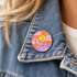 A close-up of a person wearing a round enamel pin on the collar of their blue denim jacket. The pin features a retro-style sunset illustration with pink clouds, a yellow sun, and purple flowers.