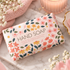 A wrapped bar of hand soap with a floral pattern sits on a marble surface surrounded by pink towels, white flowers, and a lit candle in a glass holder.