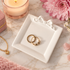 Two gold rings displayed on a white square gift-box-shaped ring holder with a bow detail, styled on a marble surface with a pink flower, candle, and jewelry accessories.