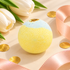 A round bath bomb with a two-tone yellow and light blue textured surface, decorated with a small gold glitter accent on top. It is styled in a flat-lay composition with white tulips, pink satin ribbon, and gold confetti on a light beige background.