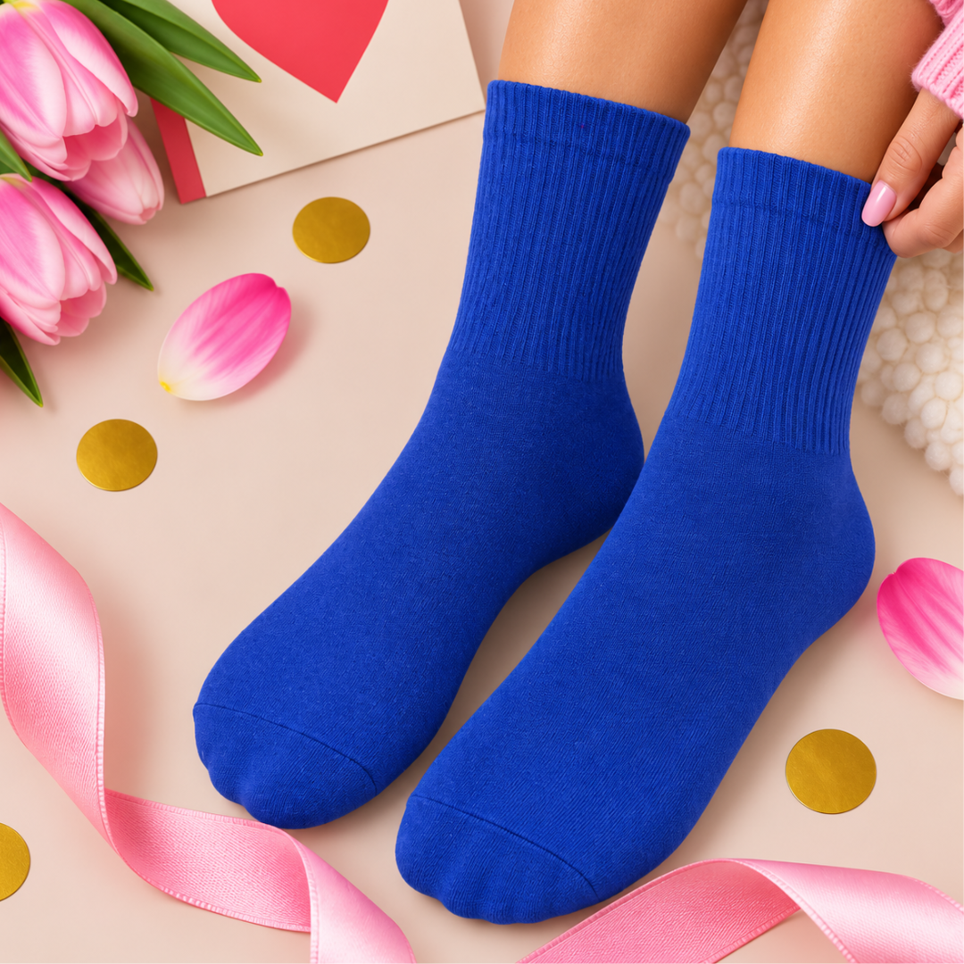 A pair of bright royal blue crew socks worn on feet, styled in a flat-lay with pink tulips, a pink ribbon, gold confetti, and a gift card on a light beige background.