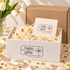 A white branded gift box filled with white paper shred and gold confetti, with a card reading &