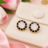 A pair of gold-tone stud earrings with black enamel and a white rose motif, displayed on a cream velvet jewelry pouch with pink tulips in the background.