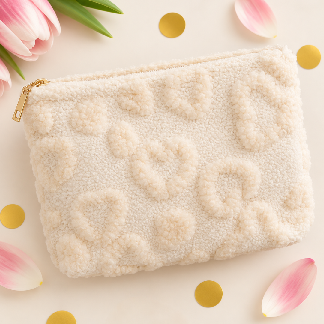 A cream-colored textured fleece pouch with raised heart patterns and a gold zipper, styled on a light beige background with pink tulips, gold confetti, and green leaves.
