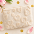 A cream-colored textured fleece pouch with raised heart patterns and a gold zipper, styled on a light beige background with pink tulips, gold confetti, and green leaves.