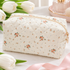 A quilted cosmetic pouch with a cream background and a delicate floral print, styled with white tulips and a pink ribbon on a light marble surface.