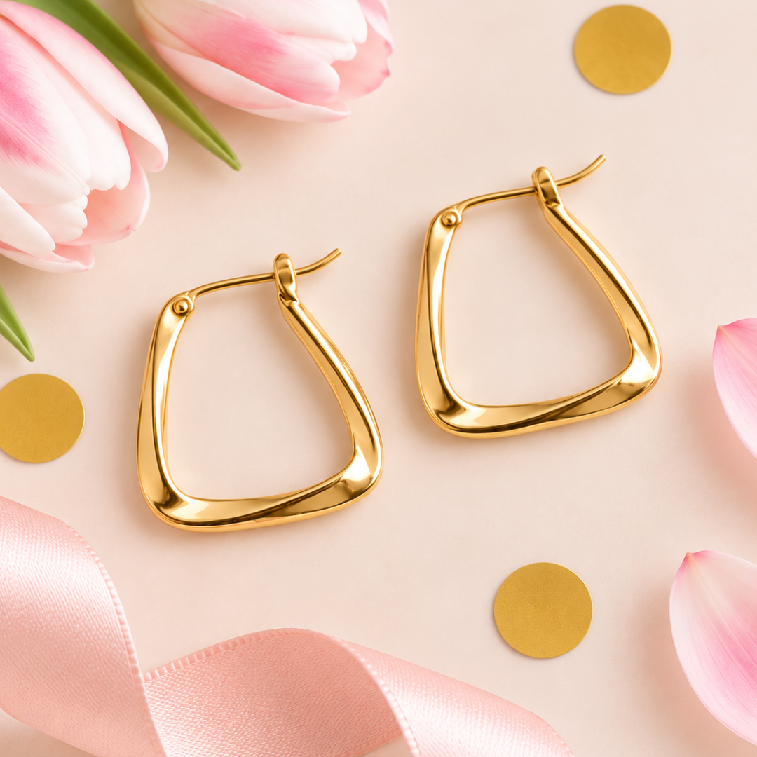 A pair of gold-toned geometric hoop earrings with a smooth, polished finish, styled on a light pink background with pink tulips, gold confetti, and a pink ribbon.