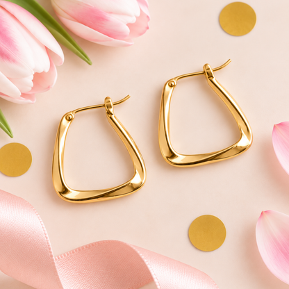A pair of gold-toned geometric hoop earrings with a smooth, polished finish, styled on a light pink background with pink tulips, gold confetti, and a pink ribbon.