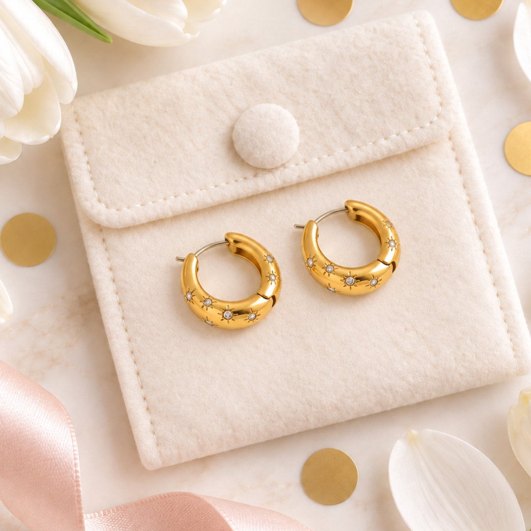 A pair of gold hoop earrings with small clear crystal accents, displayed on a cream-colored felt jewelry pouch. The pouch is styled on a marble surface with white tulips, gold confetti, and a pink satin ribbon.