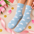 A pair of light blue crew socks with a white cloud pattern, worn on feet and styled with pink tulips, a pink ribbon, gold confetti, and a gift card.