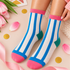 A pair of crew socks worn on feet, featuring vertical blue and white stripes with a bright pink cuff, toe, and heel, plus a green heel accent. The feet are styled on a light pink surface with tulips, gold confetti, and a pink ribbon.