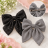 Two fabric hair bows on a light marble surface with floral props: one black bow with white polka dots and one black-and-white gingham bow. A circular inset shows the back of one bow with a metal hair clip fastener visible.