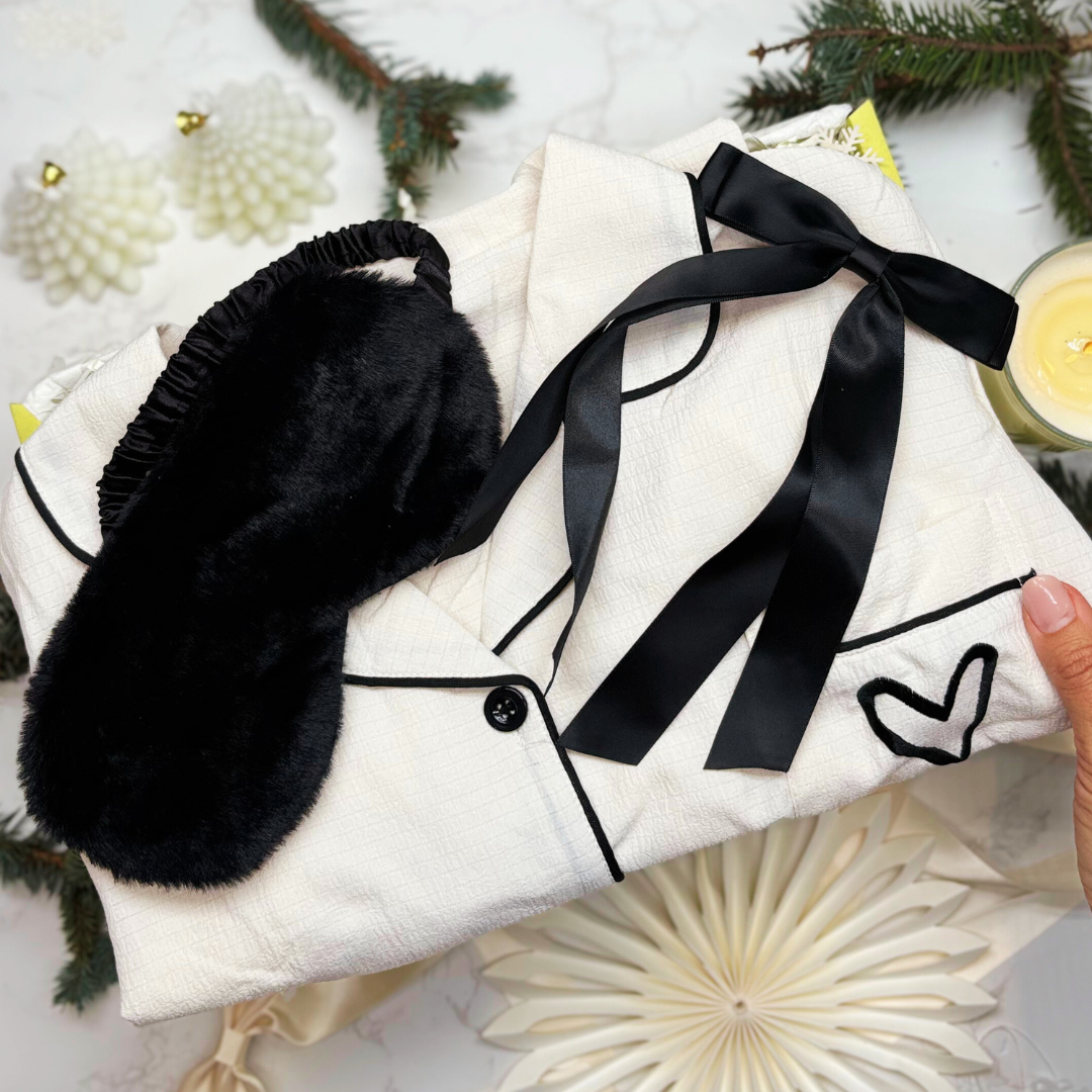 Cotton feel white pyjamas with black fluffy eye mask, and satin black hair ribbon, held against a decorative giftbox.