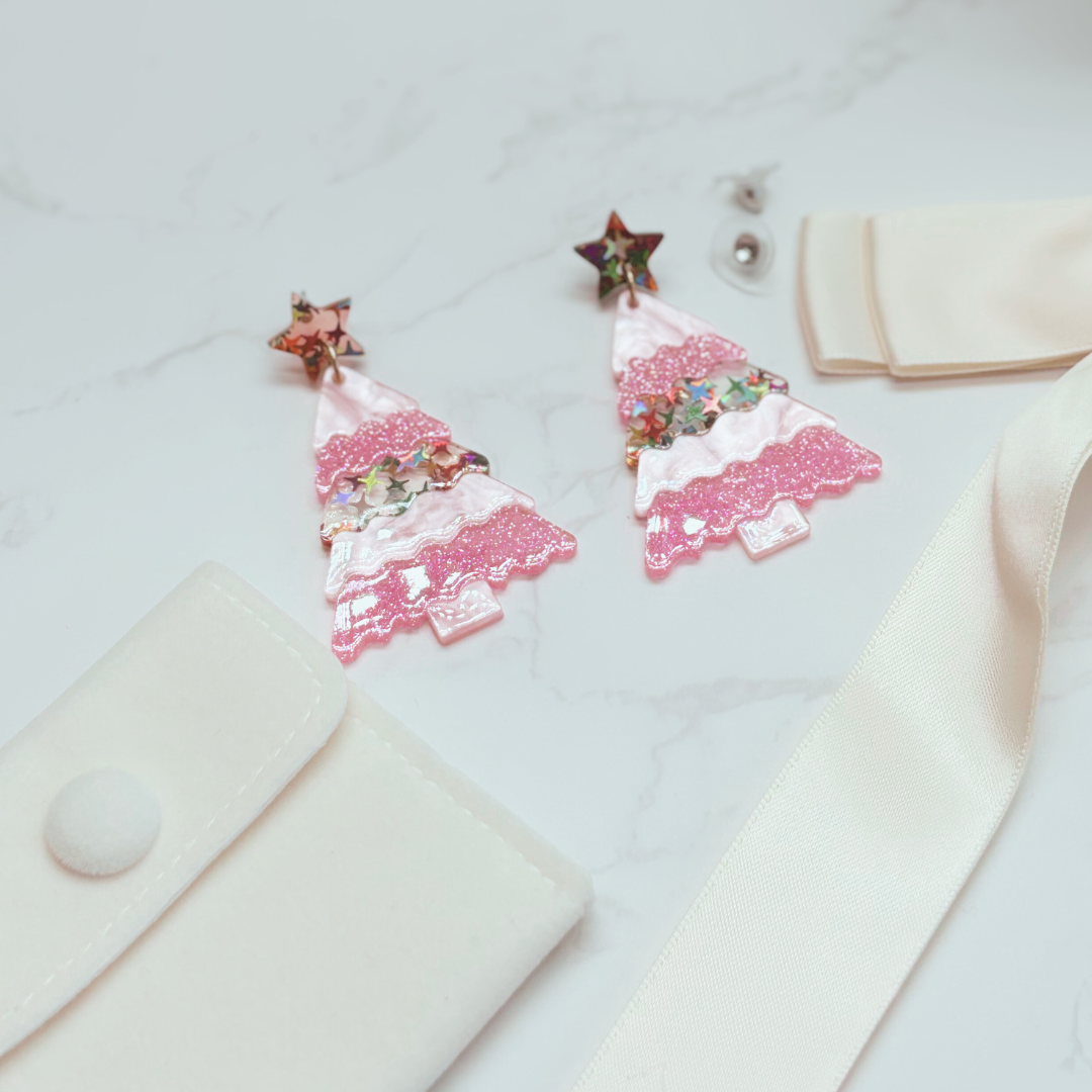 Pink Christmas tree-shaped earrings on a white surface with a beige fabric background